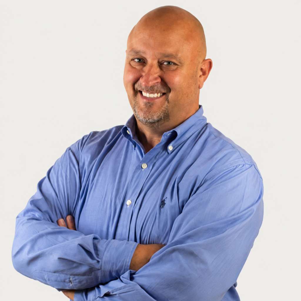 Smiling bald man in a blue button-down shirt with arms crossed against a light background.