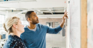 A man pointing at a whiteboard with information on it while a female colleague looks at what he is pointing at; the widening achievement and equity gaps in schools require a new initiative to bring forth action.