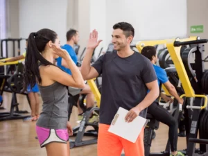 A man and a woman high-fiving in a gym. The woman wears a gray top and pink shorts. The man holds a clipboard. Humans have many microscopic bacteria such as MRSA and can be transmitted and can infect another person through sport, mainly contact sports.