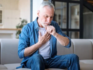 A person sitting on a couch holding his chest, coughing. RSV is an upper respiratory virus that has methods that can help control transmission like ActivePure Technology.