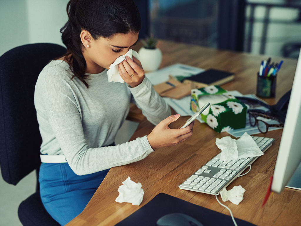 Woman at her desk blowing her nose and looking at her cell phone.