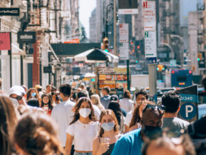 Crowded city street with pedestrians, many wearing masks, storefronts and signs in the background.