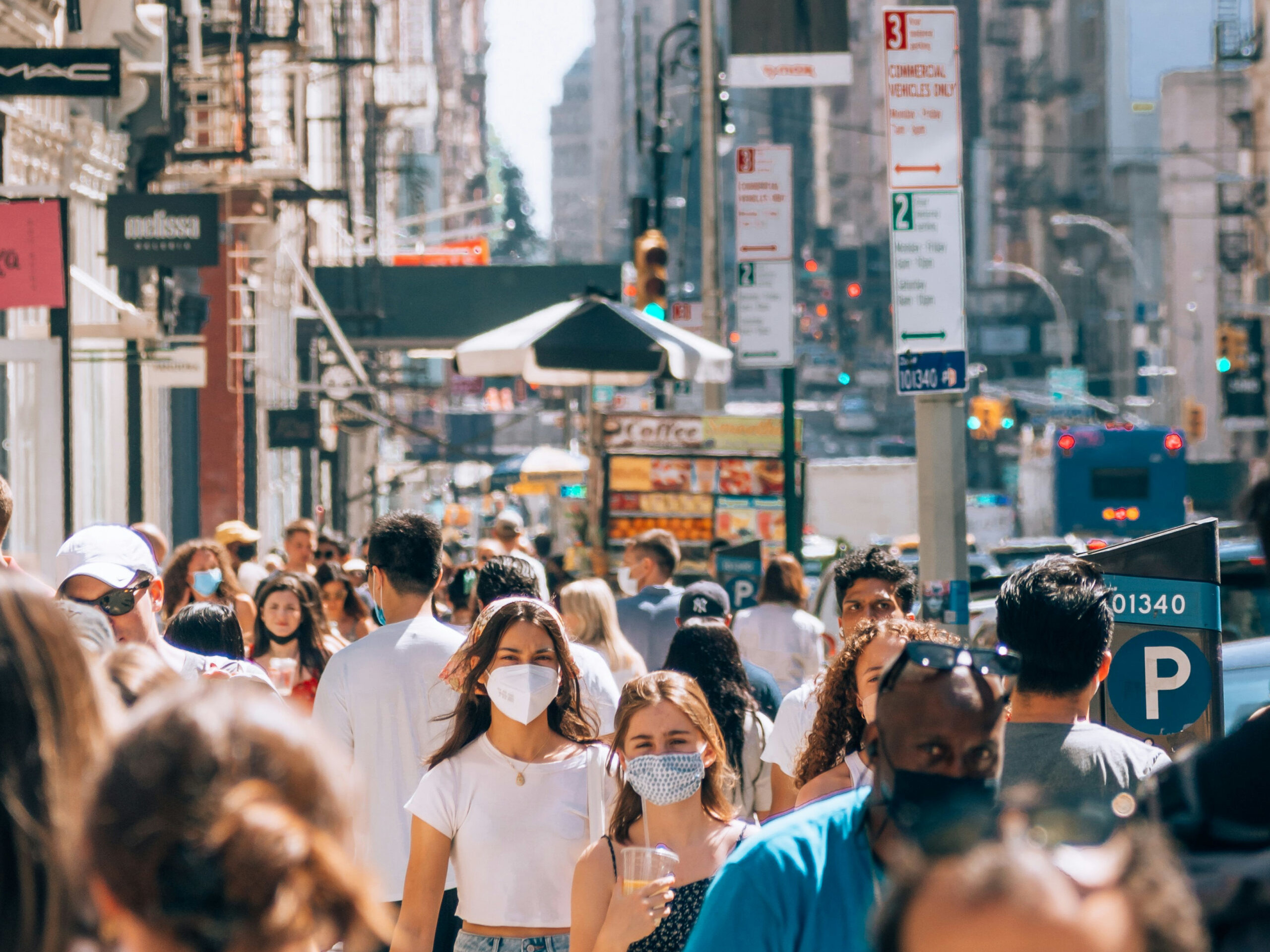 Crowded city street with pedestrians, many wearing masks, storefronts and signs in the background.