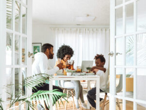 Family of four sharing a meal at a white dining table in a bright room with glass doors and plants nearby.