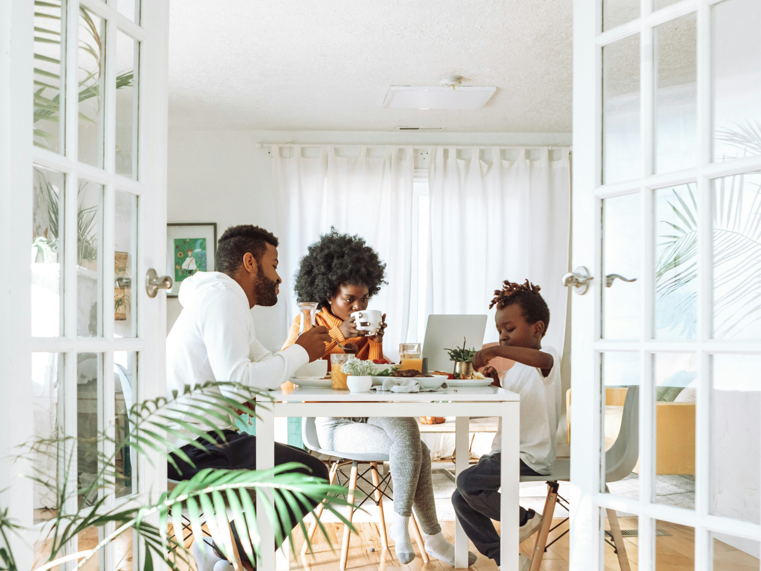 Family of four sharing a meal at a white dining table in a bright room with glass doors and plants nearby.
