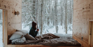 Woman sitting on bad in front of window with snow covering forest landscape