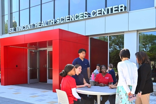 Group of diverse people sit around a round table outside St. Vincent Health Sciences Center near a red entrance.